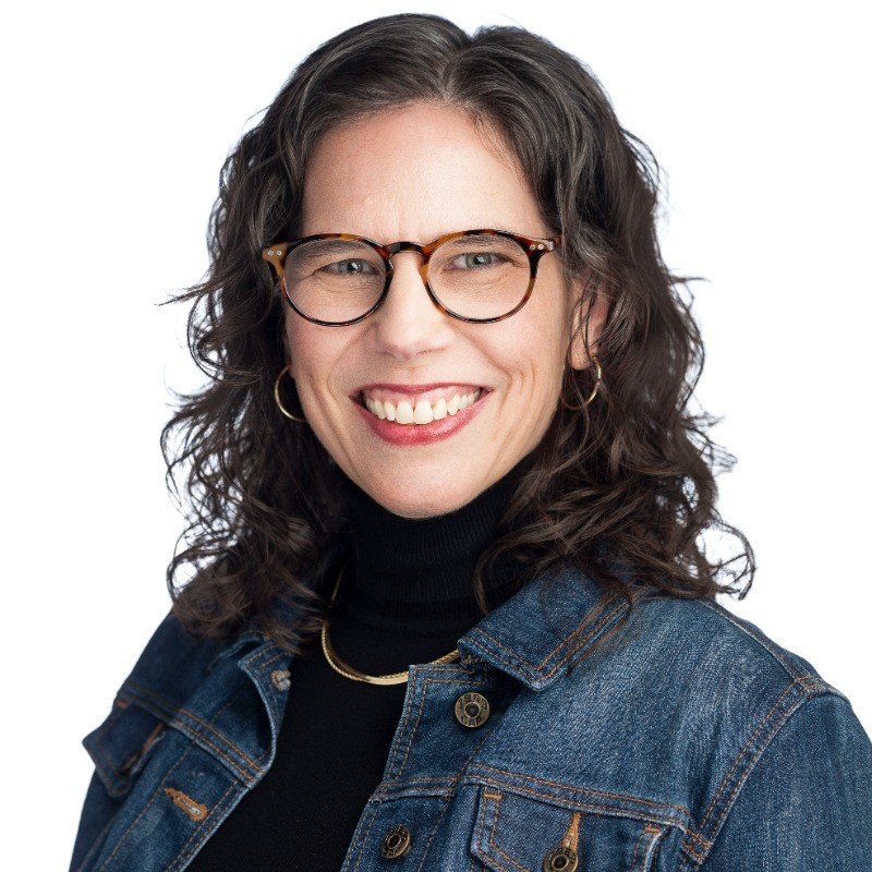 Woman with wavy brown hair, wearing glasses, a denim jacket, black turtleneck, and gold jewelry, smiling at the camera against a white background.